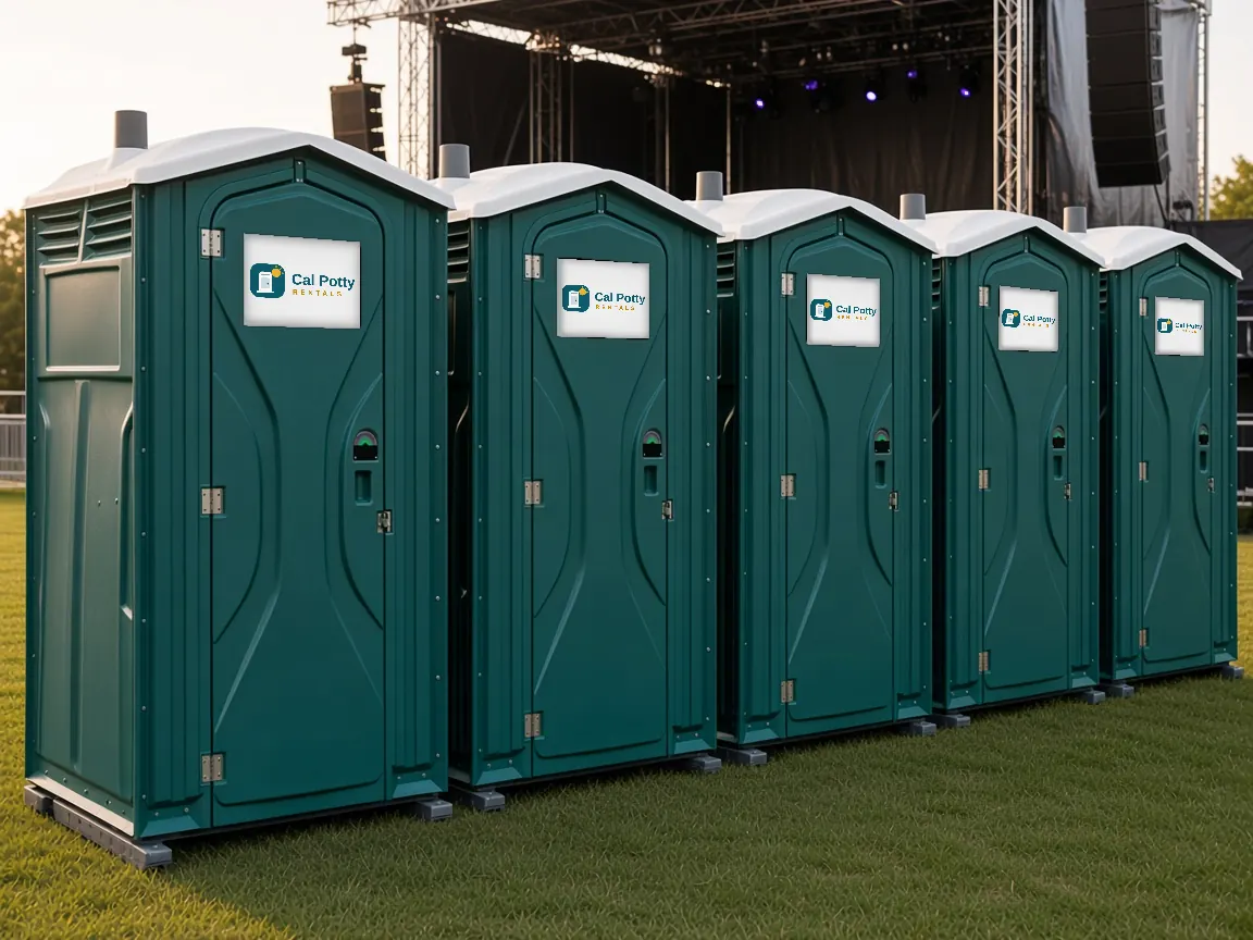 Five teal portable toilets in a row on a lawn near a concert stage